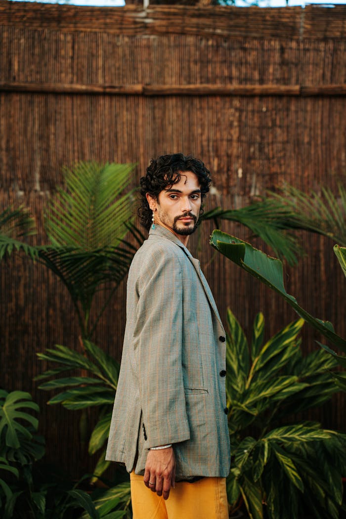A fashionable adult male posing confidently outdoors surrounded by lush green tropical plants.