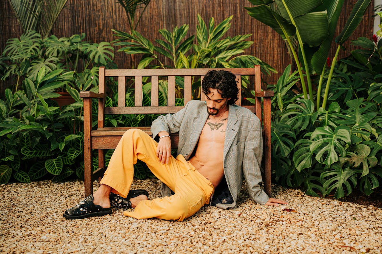 A fashionable man relaxes on a wooden bench surrounded by lush tropical plants in a garden setting.